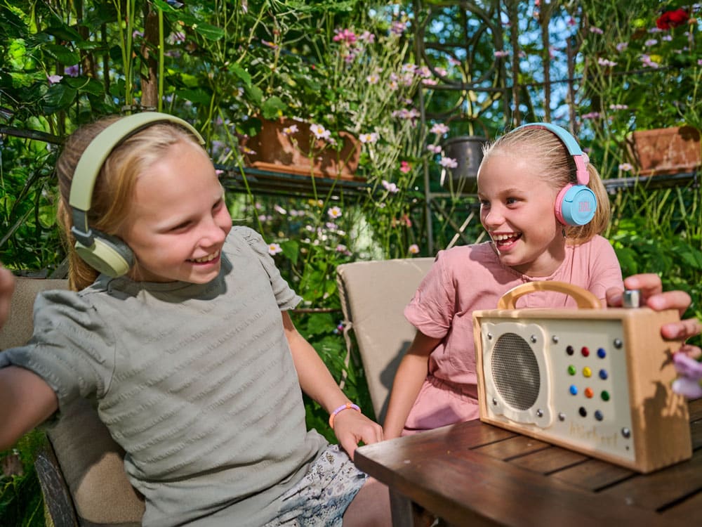 hörbert steht auf einem Gartentisch, und zwei Mädchen lauschen mit Bluetooth-Kopfhörern