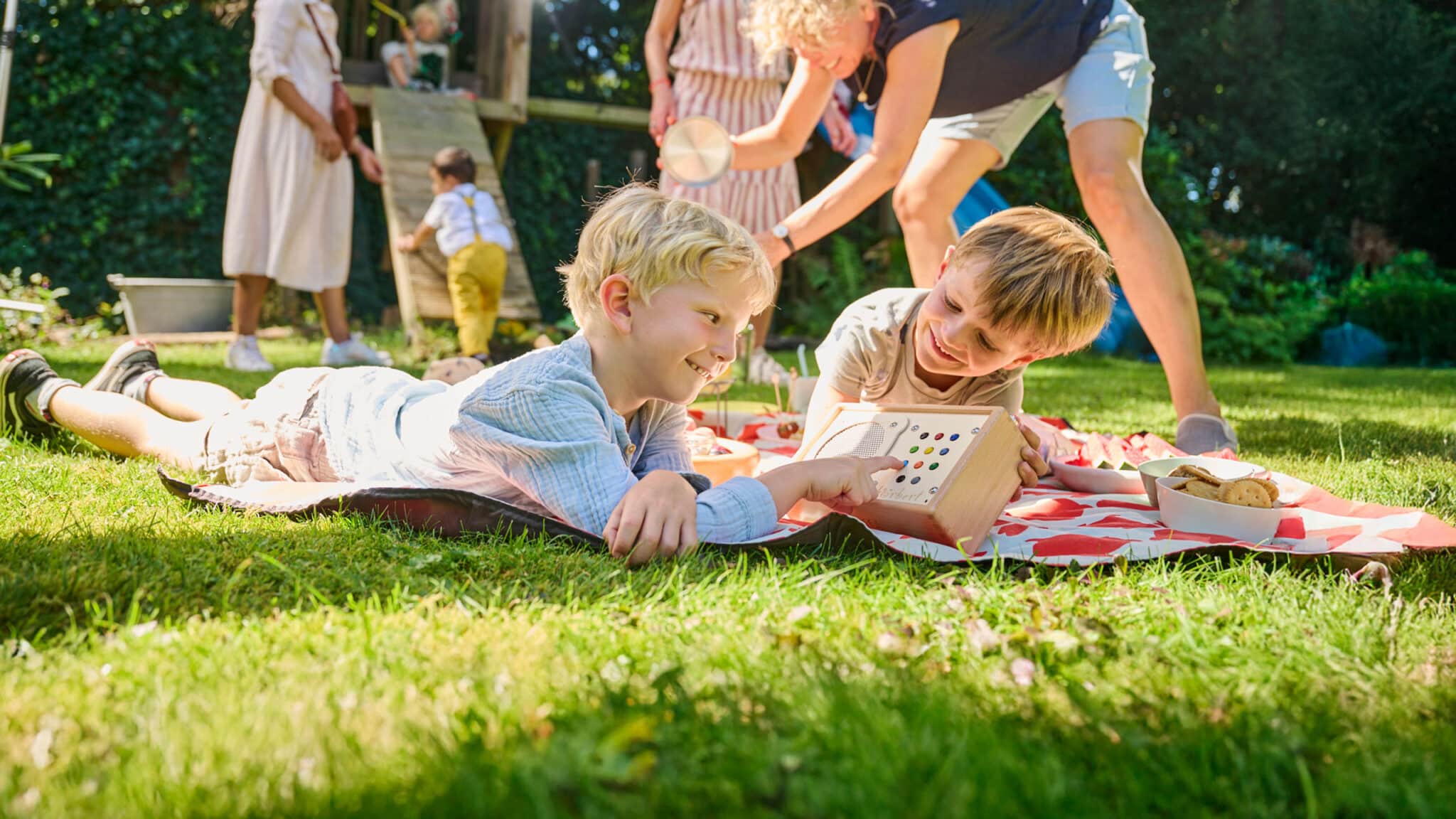 Zwei Jungen hören auf der Picknickdecke hörbert zu, während im Hintergrund andere Kinder spielen