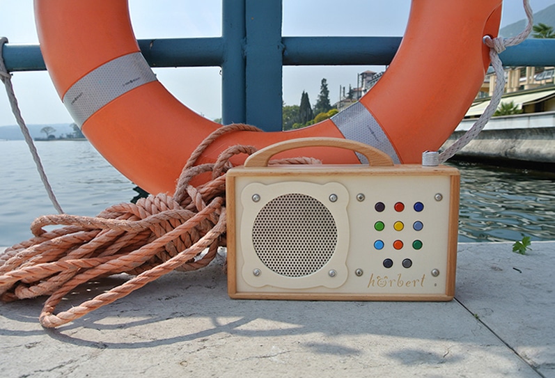 In diesem Bild sieht man einen hörbert Musikspieler mit 9 Playlists, der auf einem Landungssteg eines Gewässers steht. Im Hintergrund ist ein orange-weiss gestreifter Rettungsring zu sehen und Taue.